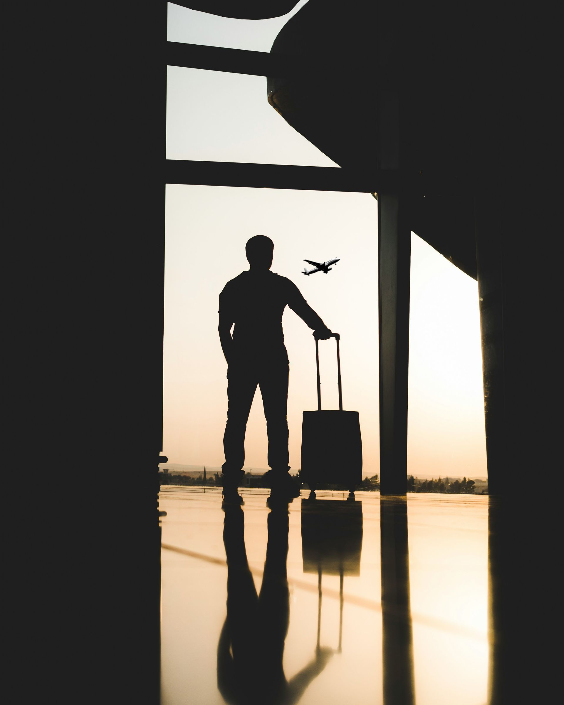 Silhouette of a person with a suitcase in front of a window, watching a plane take off, airport setting.