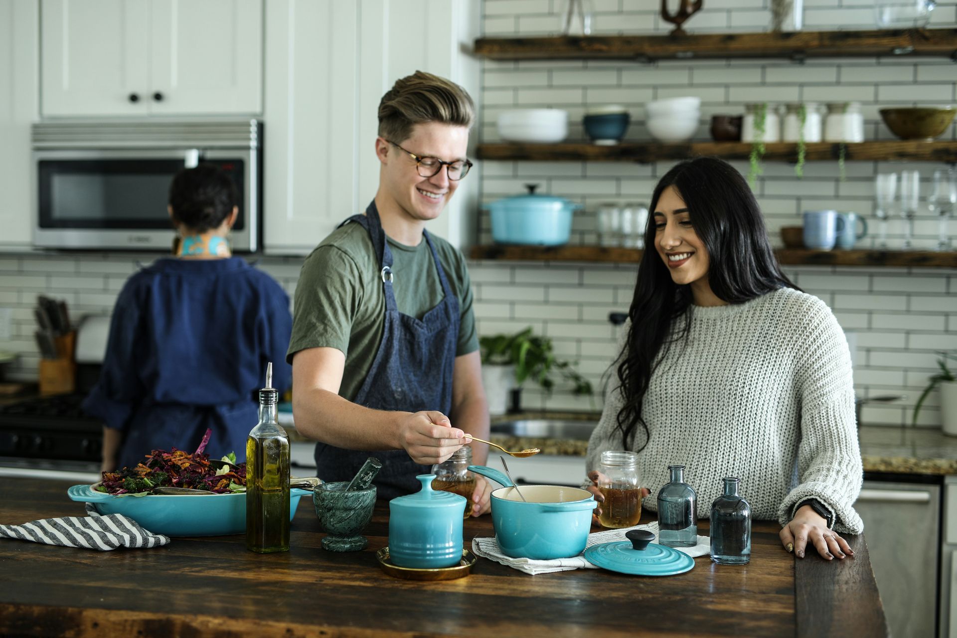 Couple in kitchen, smiling, cooking. Wooden counter, blue cookware, person in blue behind.