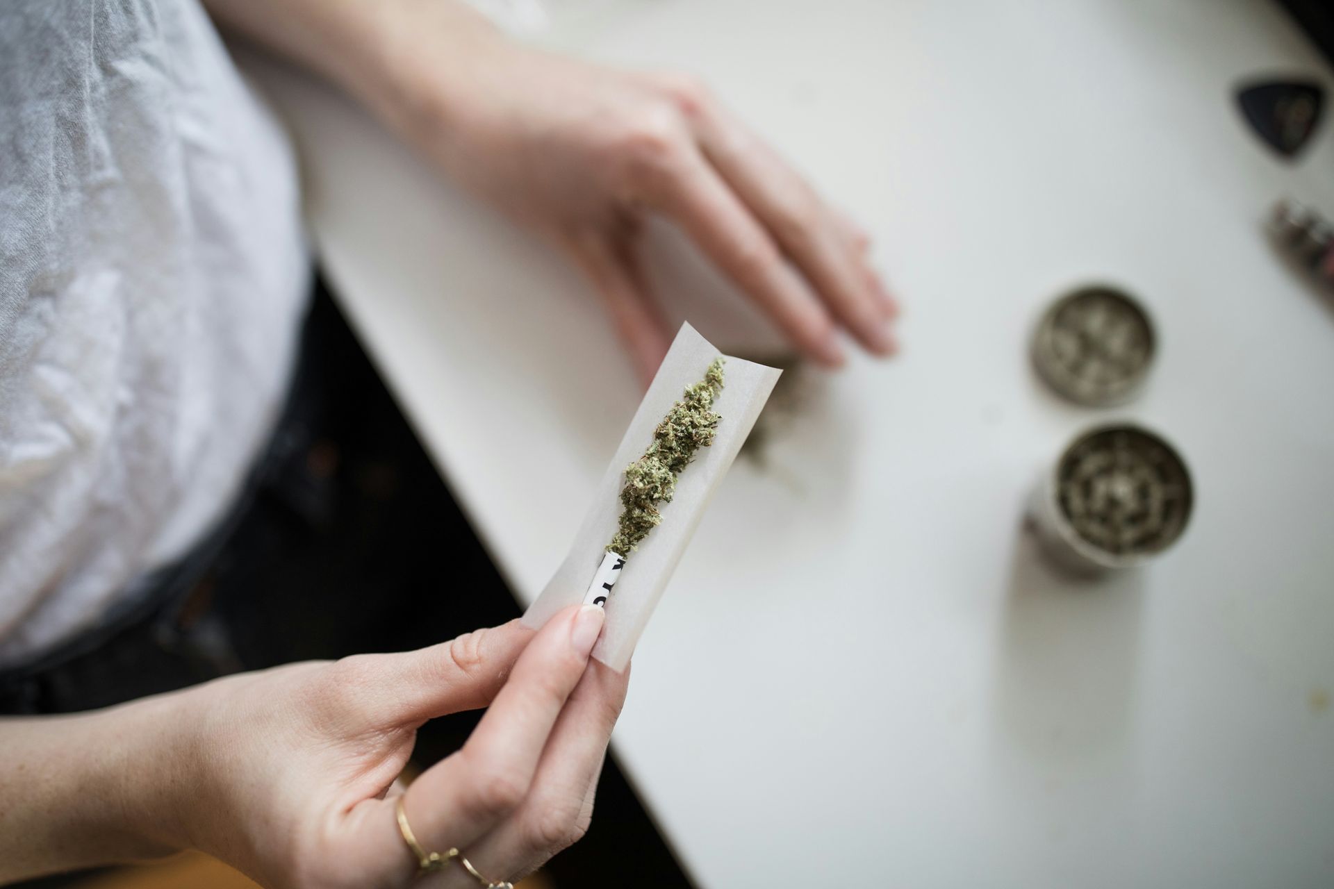 Hands rolling a marijuana joint on a white surface, with two grinders nearby.