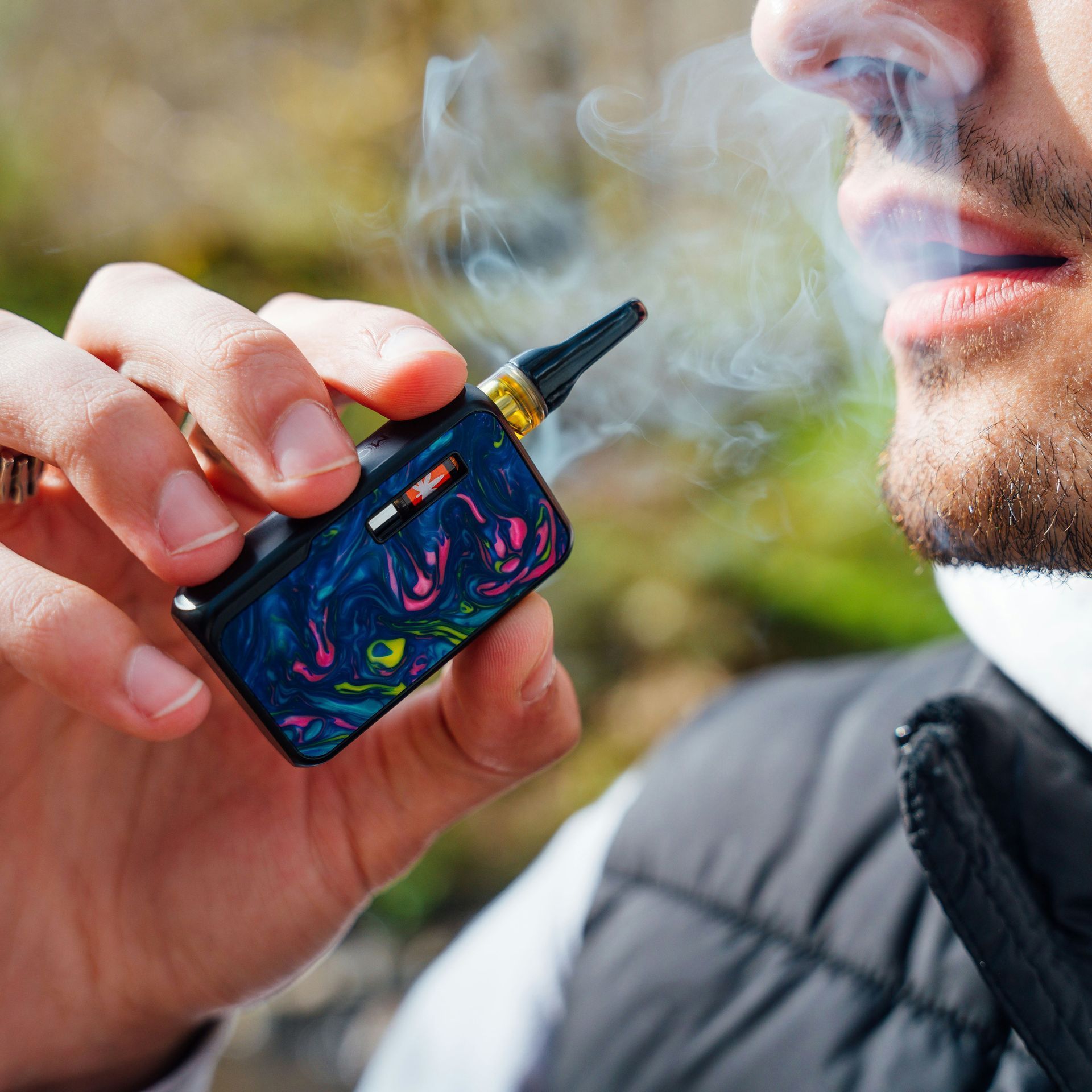 Man exhales vapor from a colorful vape device held in his hand.