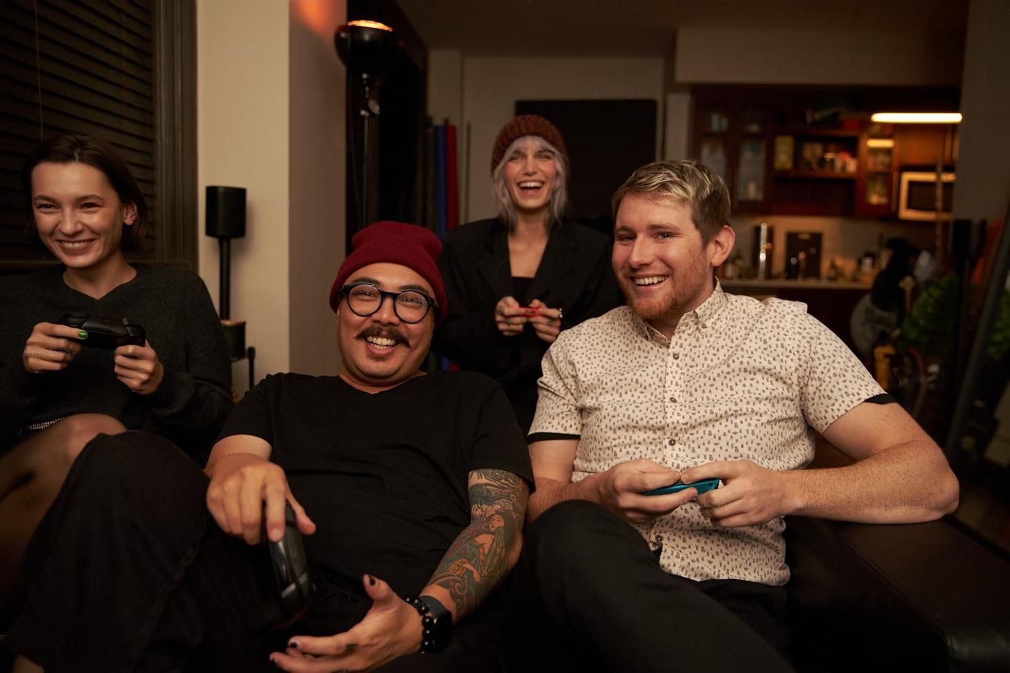 Four people playing video games on a couch, laughing. Living room setting.