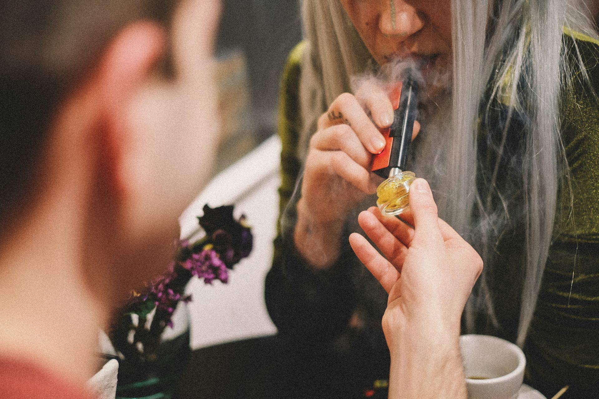 Person lighting a small glass pipe for another person. Indoor setting, smoky.