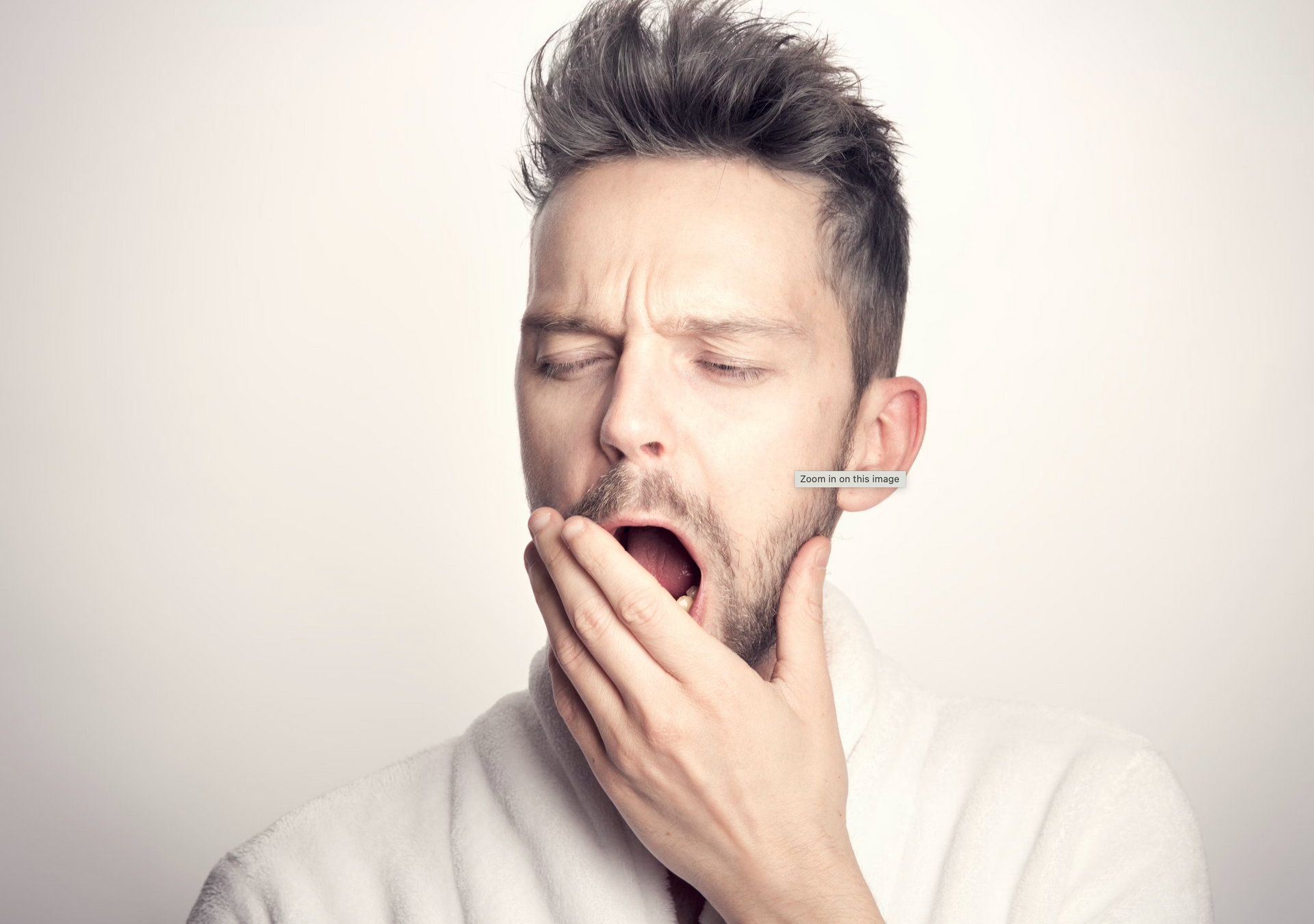 Man yawning, eyes closed, hand covering mouth, wearing a white robe, metal earring.