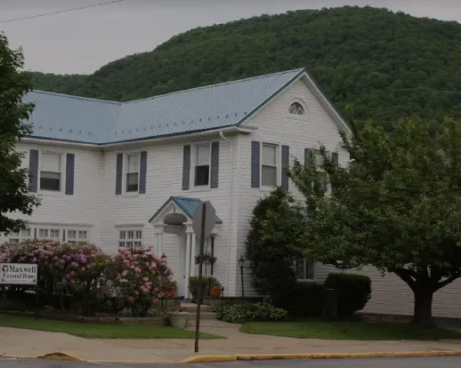 White house with blue-gray roof and a flowering bush, set against a green hillside.