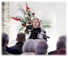 Speaker at podium with microphone, addressing an audience in a bright room with floral arrangement behind her.