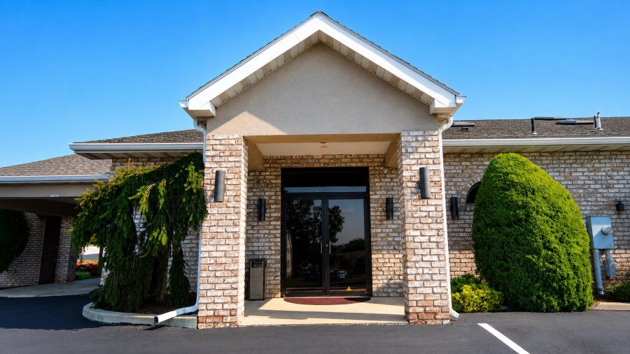 Brick and stone funeral home entrance with glass doors, landscaped shrubs, blue sky, Funeral Homes in Red Lion, PA