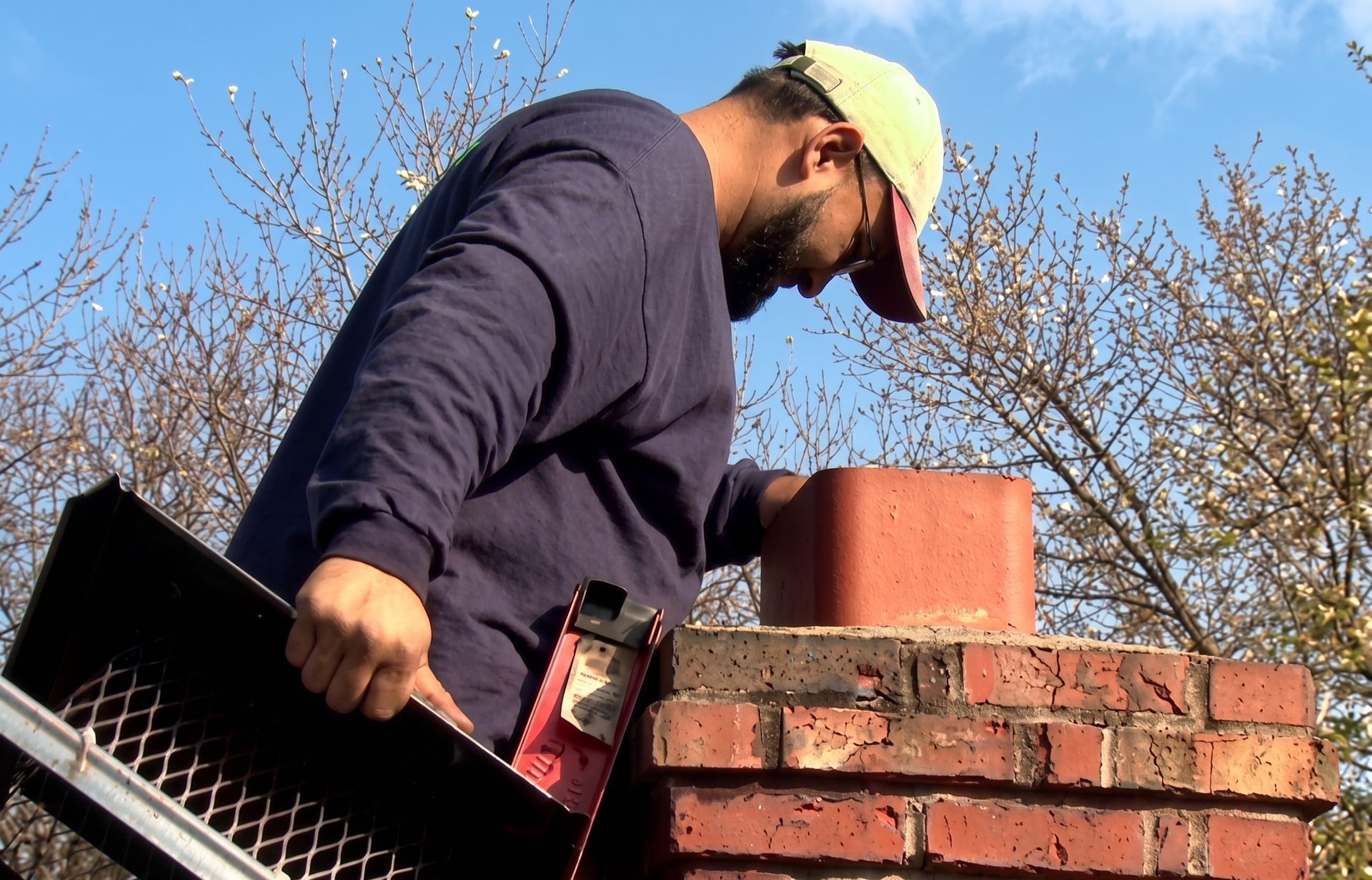 Worker on a brick chimney roof under a blue sky, using a trowel and tool bag.