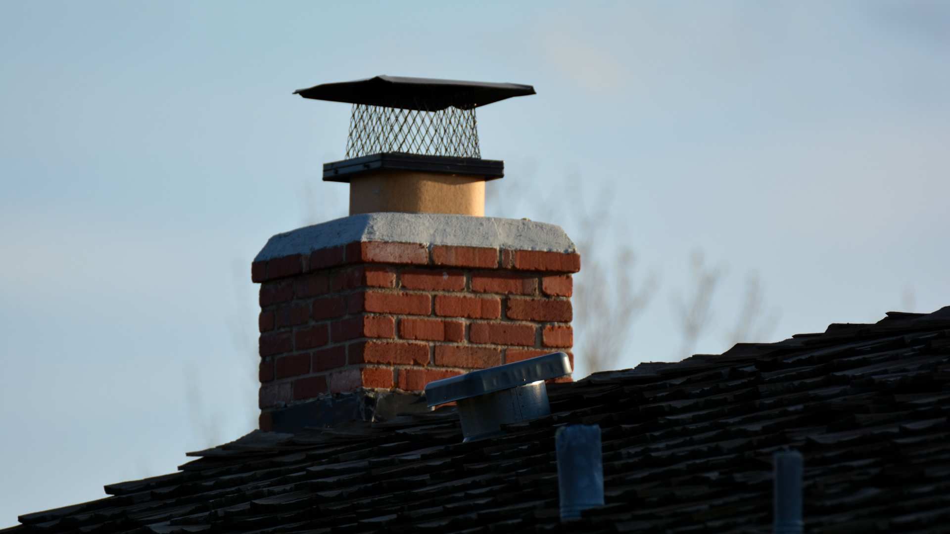 Brick chimney with a black cap on a shingled roof against a clear sky