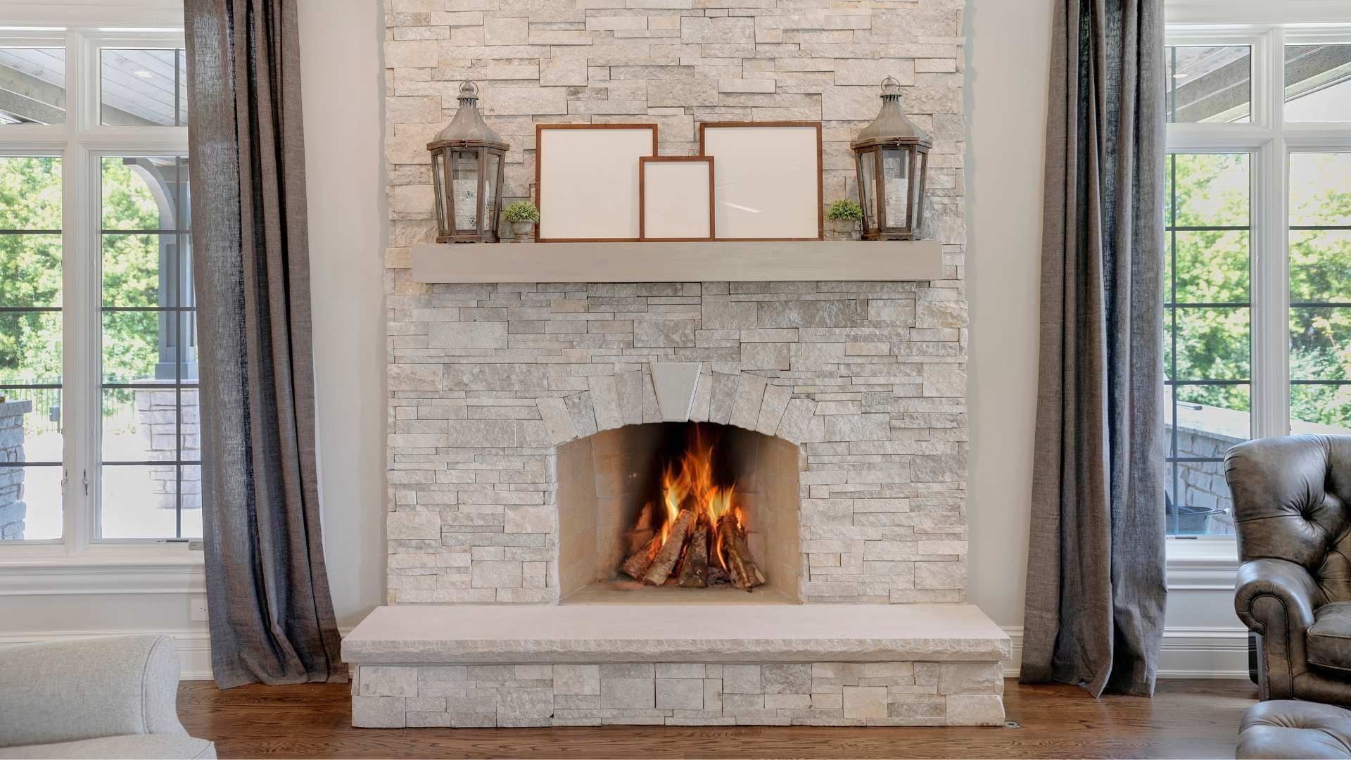 Bright living room fireplace with lit fire, white stone surround, mantel decor, and tall windows on both sides