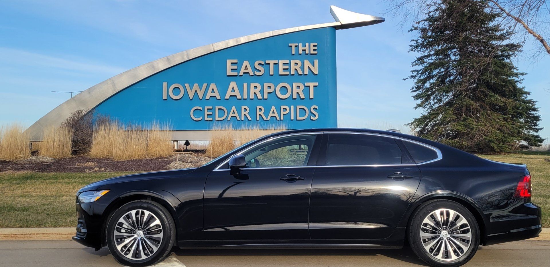 A black Volvo S90 sedan parked in front of a large blue sign reading