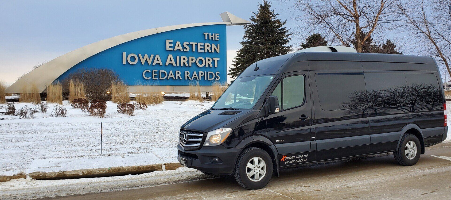A black Mercedes Sprinter Van is parked in front of a large sign reading 'The Eastern Iowa Airport Cedar Rapids' in a snowy landscape, ready to serve as the Cedar Rapids airport shuttle.