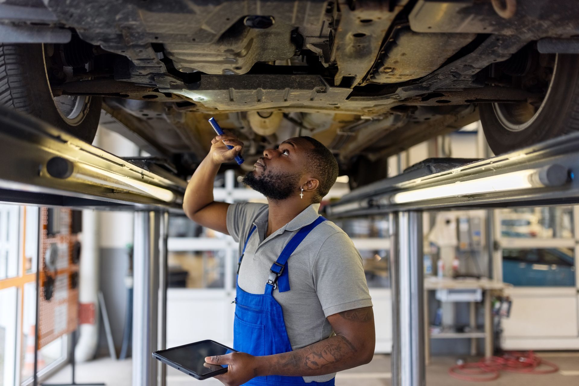 A mechanic checks the car's underside with a flashlight and tablet.