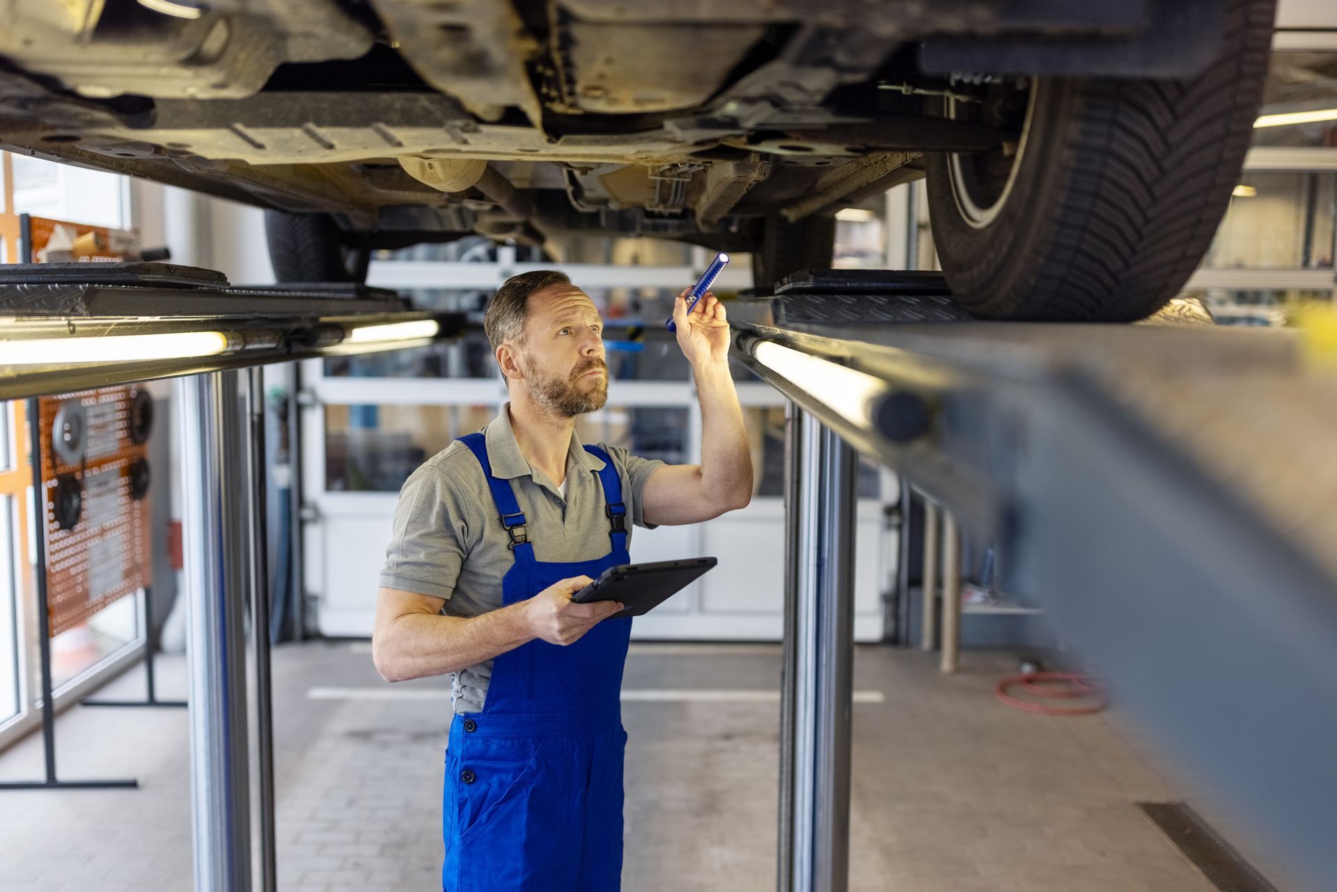 A mechanic inspects the car underside with a tablet and pen in the garage.