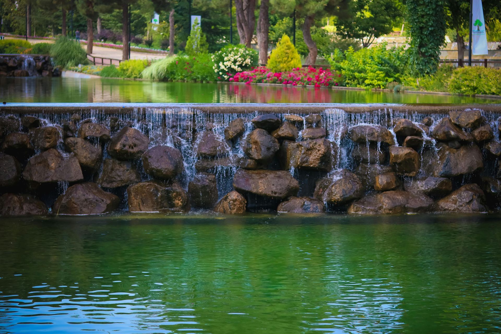 Waterfall cascading into a green pond at a park, surrounded by lush greenery and flowers.