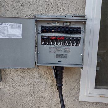 Gray electrical panel mounted outdoors next to a window. The panel door is open, revealing black and red circuit breakers.