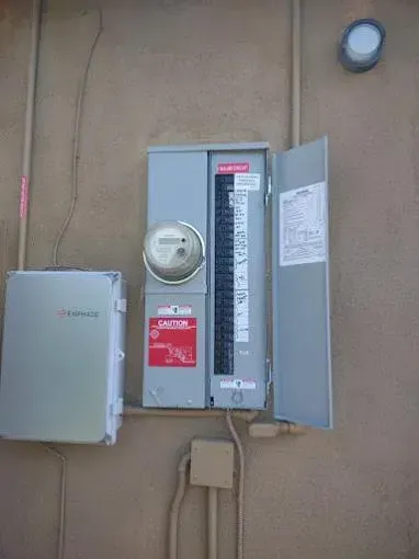 An outdoor electrical panel with a meter, circuit breakers, and open door against a stucco wall.