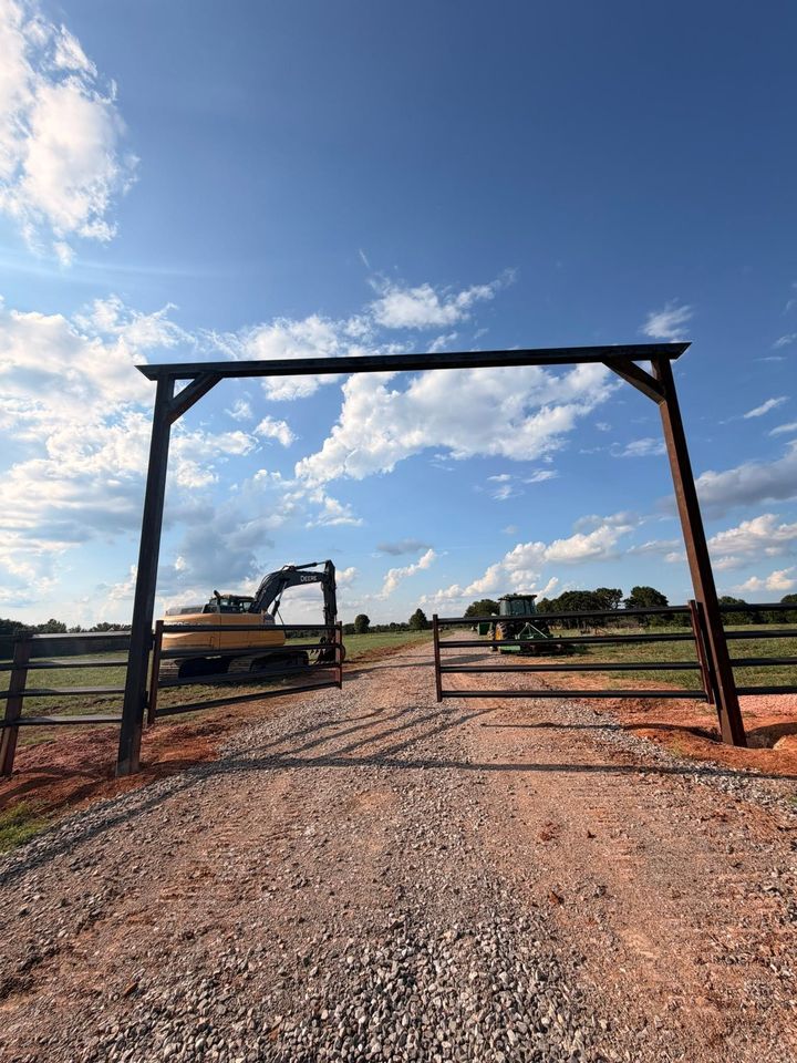 Rustic ranch entrance with a wooden gate, gravel path, and excavator under a blue sky with clouds