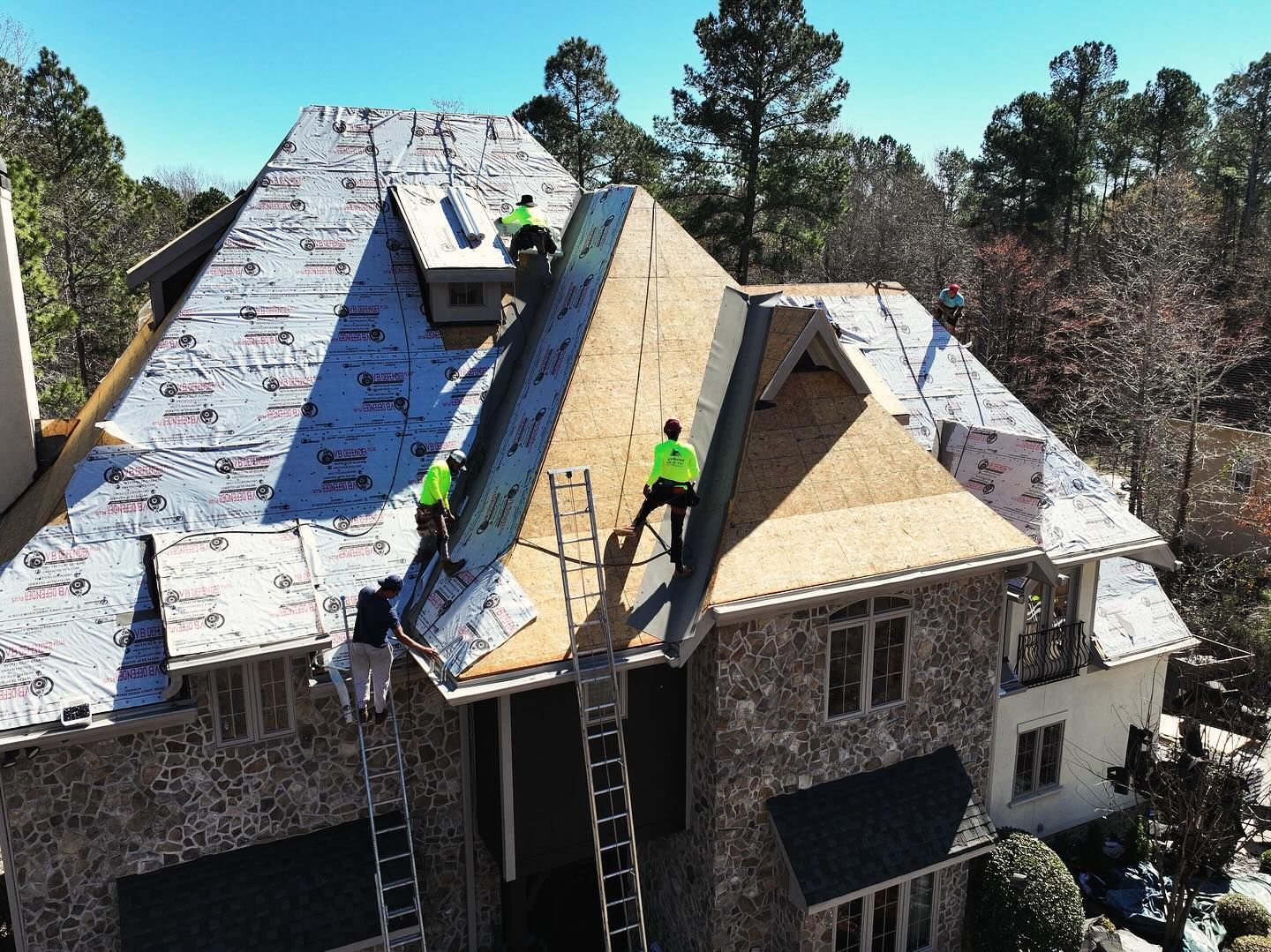 Roofers installing shingles on a steep house roof, with ladders and blue underlayment visible.