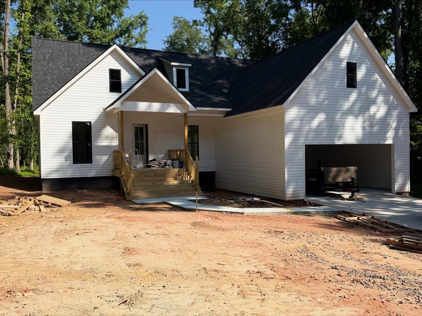 White house with black roof and attached garage under construction on a dirt lot, viewed from the front