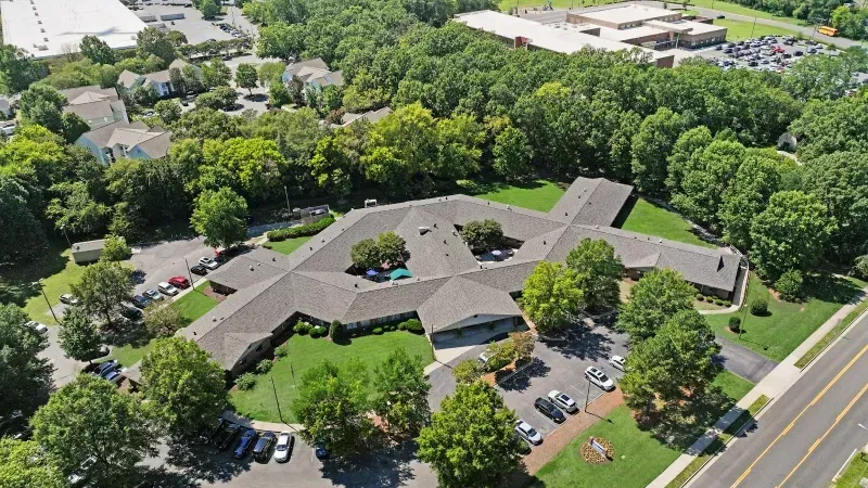 Aerial view of a long, gray-roofed office building surrounded by trees and a parking lot beside a road.