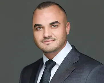 Studio headshot of a man in a gray suit, white shirt, and black tie against a gray background