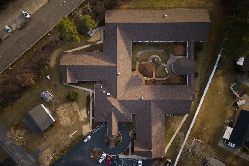 Aerial view of a brown-roofed house with a courtyard and surrounding driveway, trees, and parked cars.
