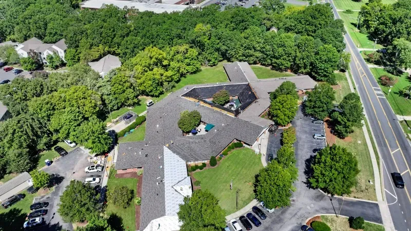Aerial view of a building with a parking lot beside a tree-lined road and dense greenery