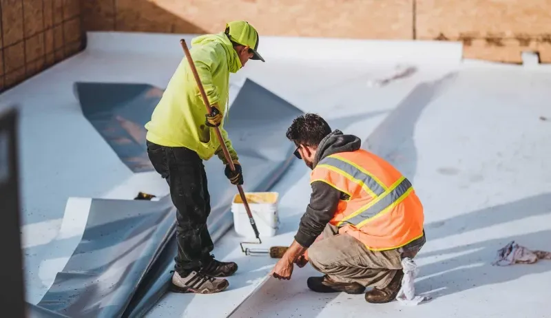 Two workers in safety vests inspect and repair a rooftop surface with tools.