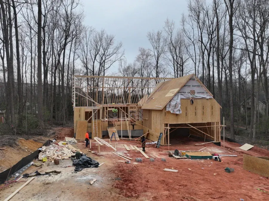 House under construction with wooden framing, workers, and building materials on a red dirt lot by bare trees