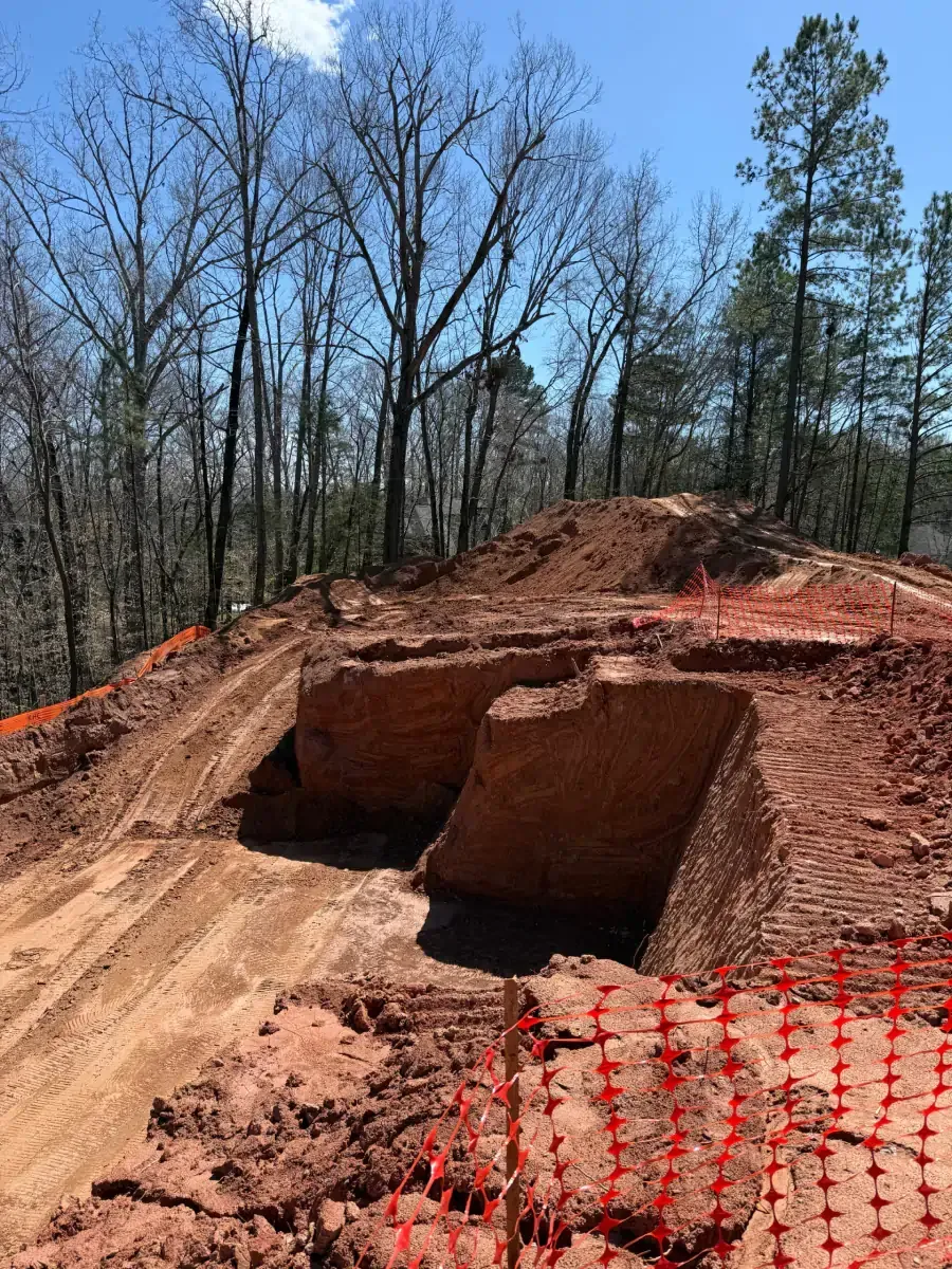 Red dirt excavation pit with orange safety fencing in a wooded area under a blue sky