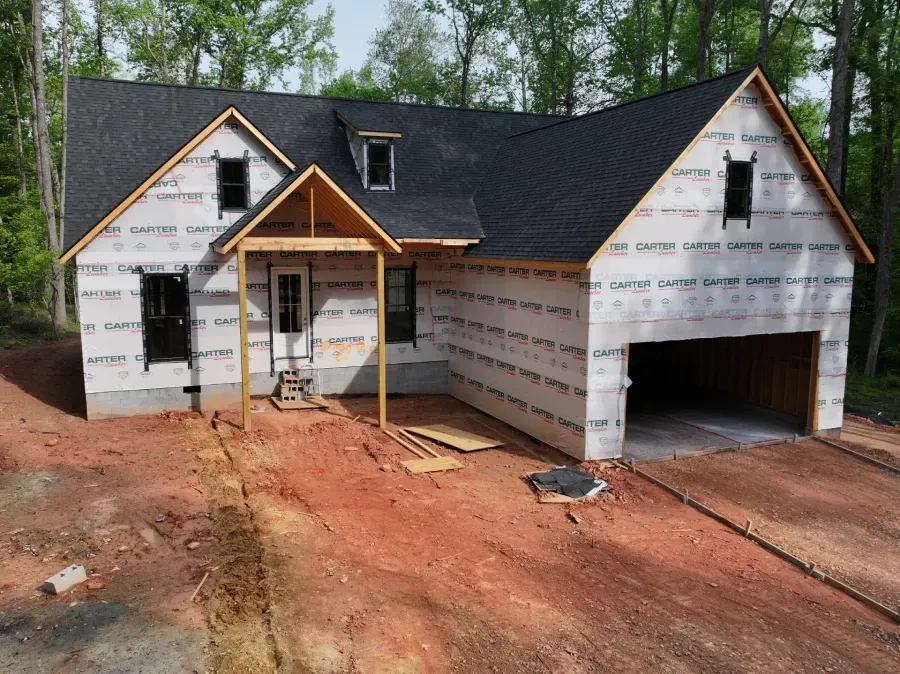 Unfinished house exterior with white wrap, black roof, garage, and dirt driveway under construction