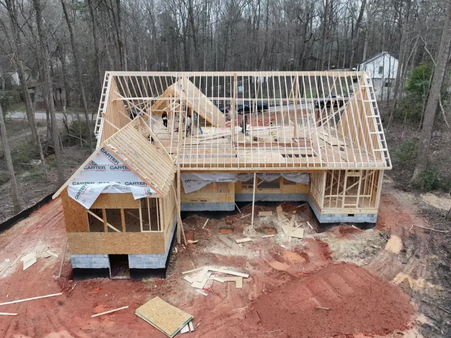 Partially framed house under construction on a red dirt lot surrounded by bare trees
