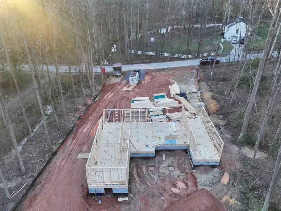 Aerial view of a wooden house under construction on a red dirt lot, surrounded by trees and a road.