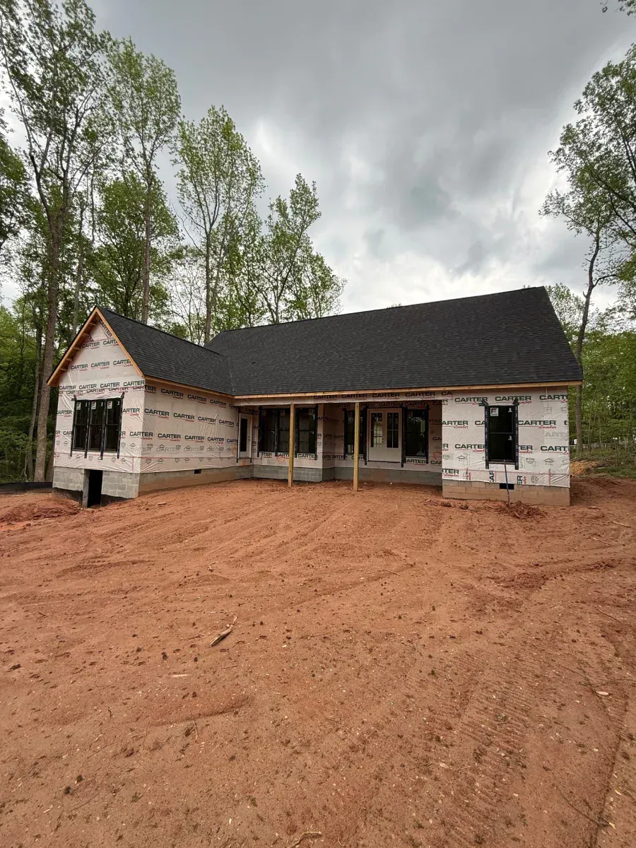 Small unfinished house with white siding and dark roof on a muddy lot, surrounded by trees under cloudy sky