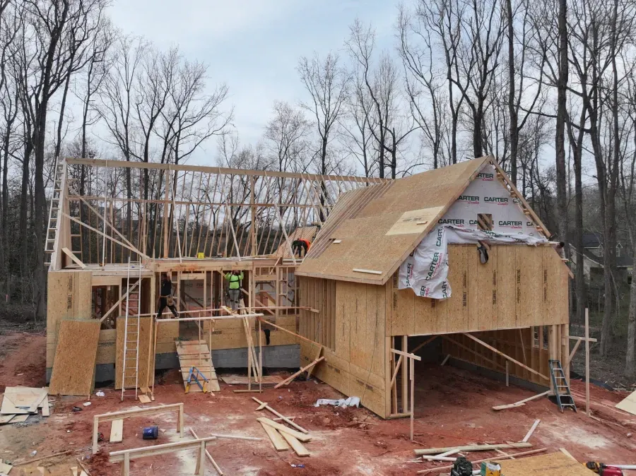 Partially built wooden house with exposed framing and roof under construction in a wooded area