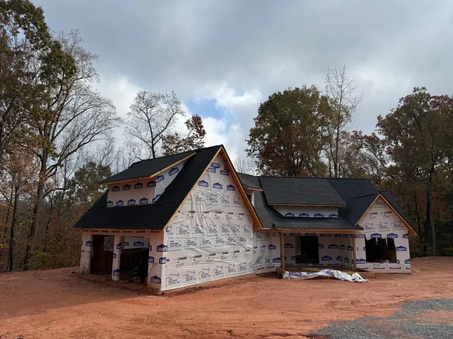 Partially constructed house with white house wrap, black roof, and red dirt clearing under cloudy sky