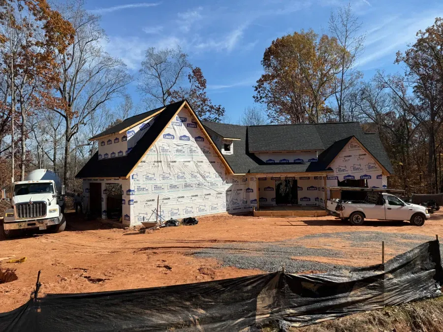 Partially built house with white siding, trucks, and bare trees on a dirt lot