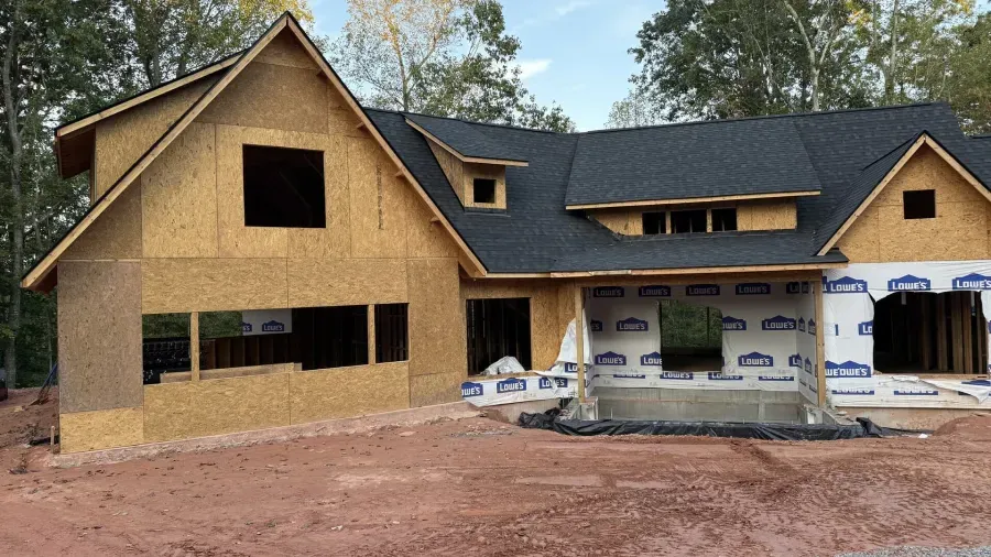 Partially built house with tan stucco exterior, black roof, and exposed framing in a wooded area