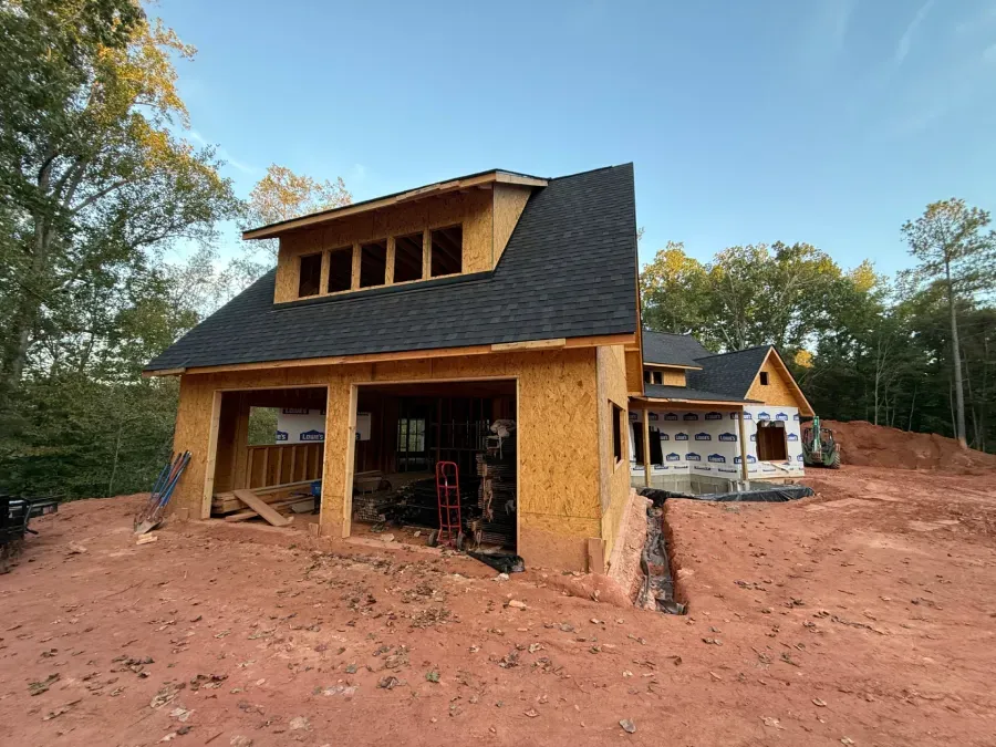 Partially built house with orange walls and dark roof on a red dirt lot surrounded by trees