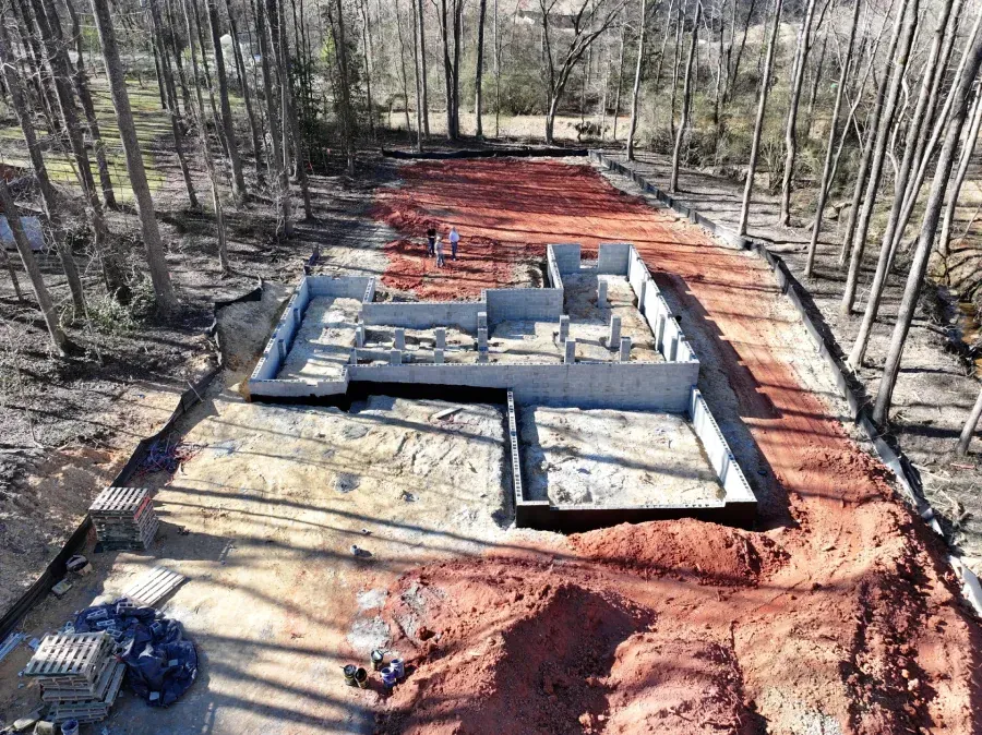 Aerial view of a foundation under construction in a wooded area with red dirt and concrete walls.