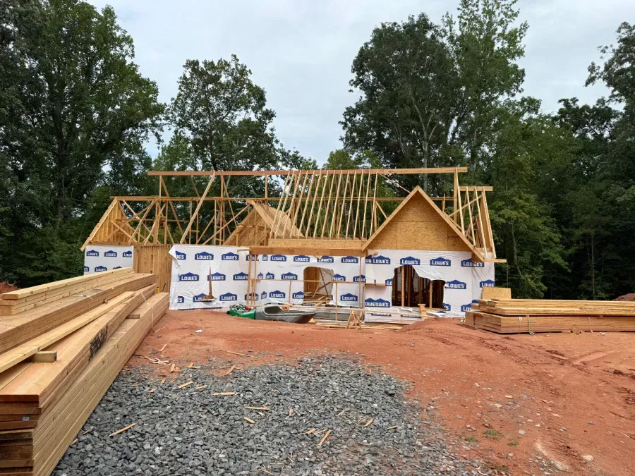 Partially built house with exposed roof trusses, lumber stacks, and house wrap on a dirt lot under cloudy sky