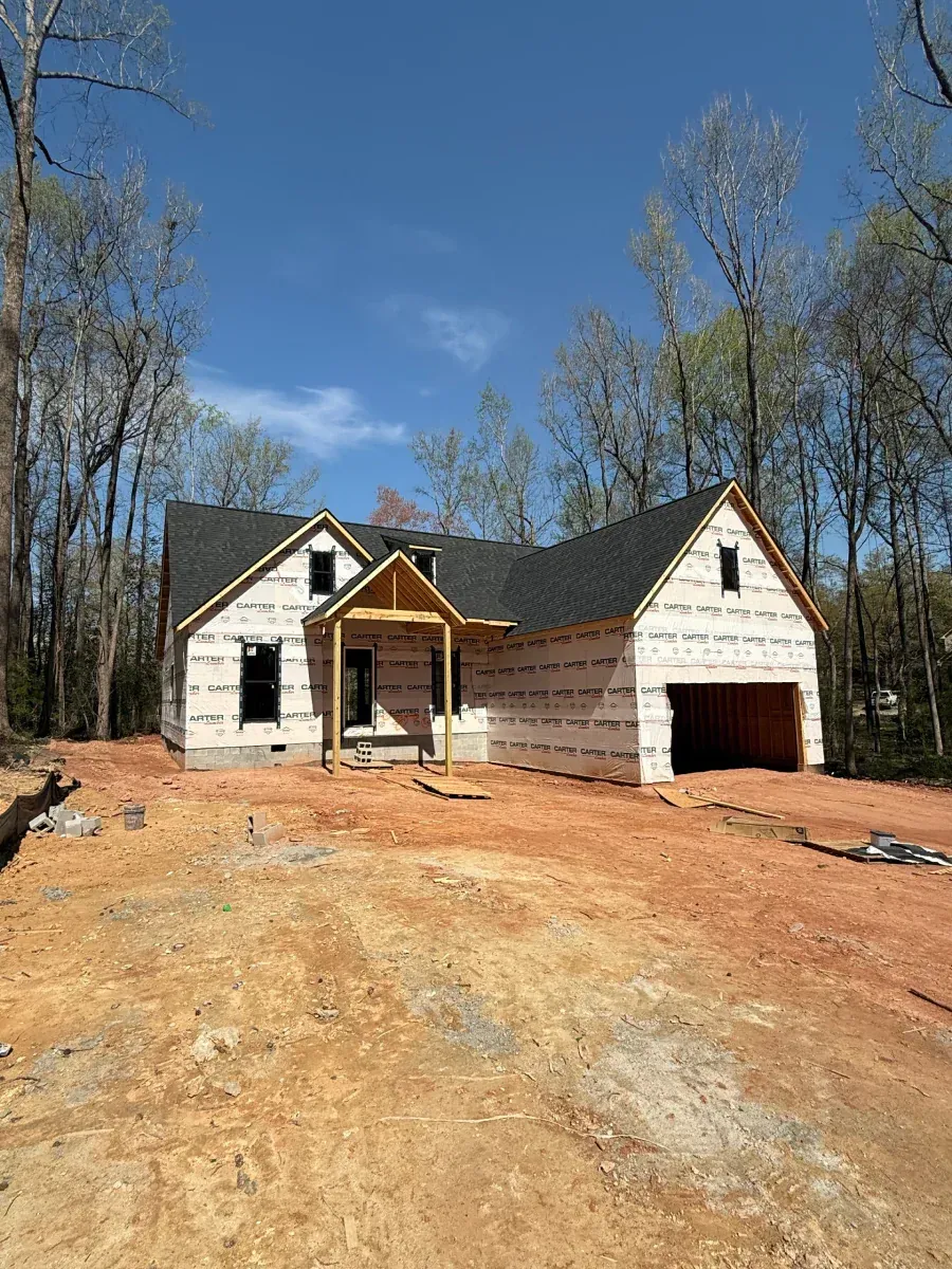 Partially built house with white siding and a garage, set on a dirt lot with bare trees and blue sky.