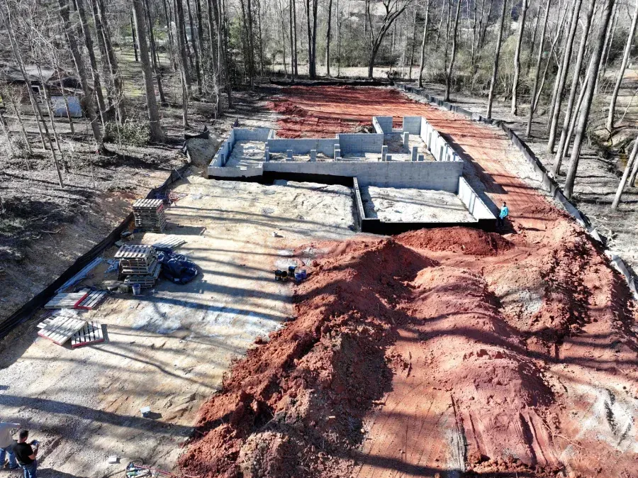 Aerial view of a house foundation under construction with red dirt and wooded surroundings