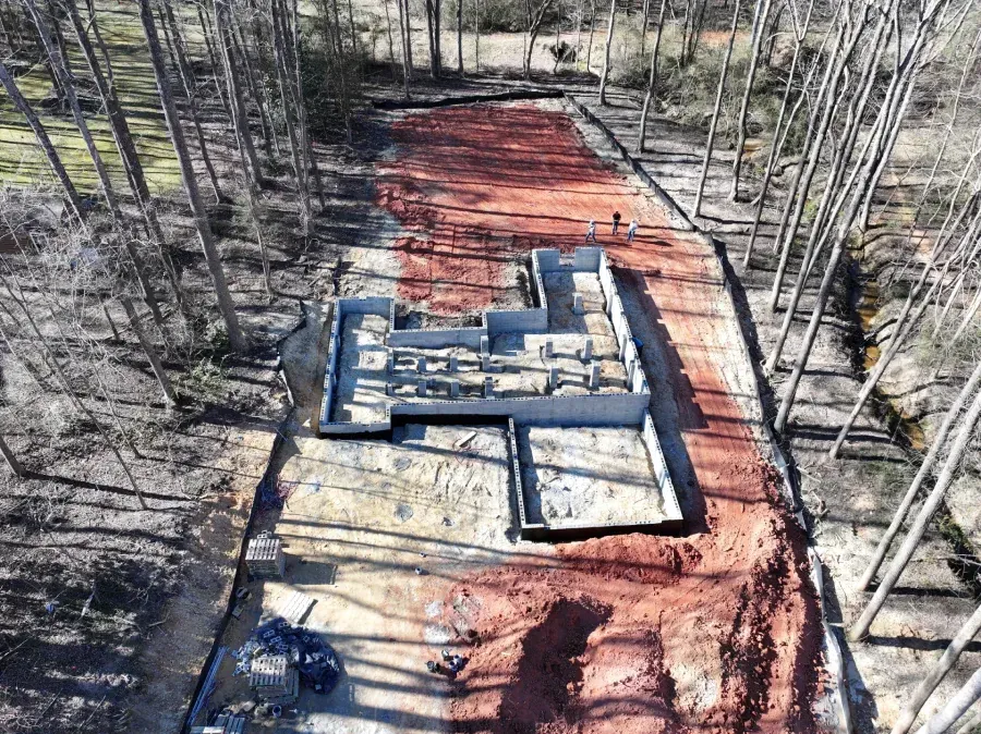Aerial view of a house foundation under construction in a wooded area, with red dirt access paths.