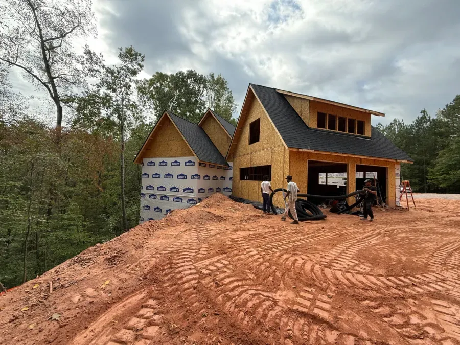 Two-story wooden house under construction on a muddy lot, with workers and forest trees nearby