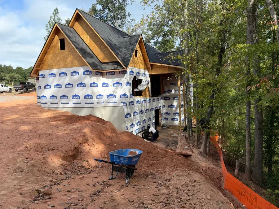 House under construction on a dirt slope, wrapped in white housewrap, with a blue wheelbarrow in front.