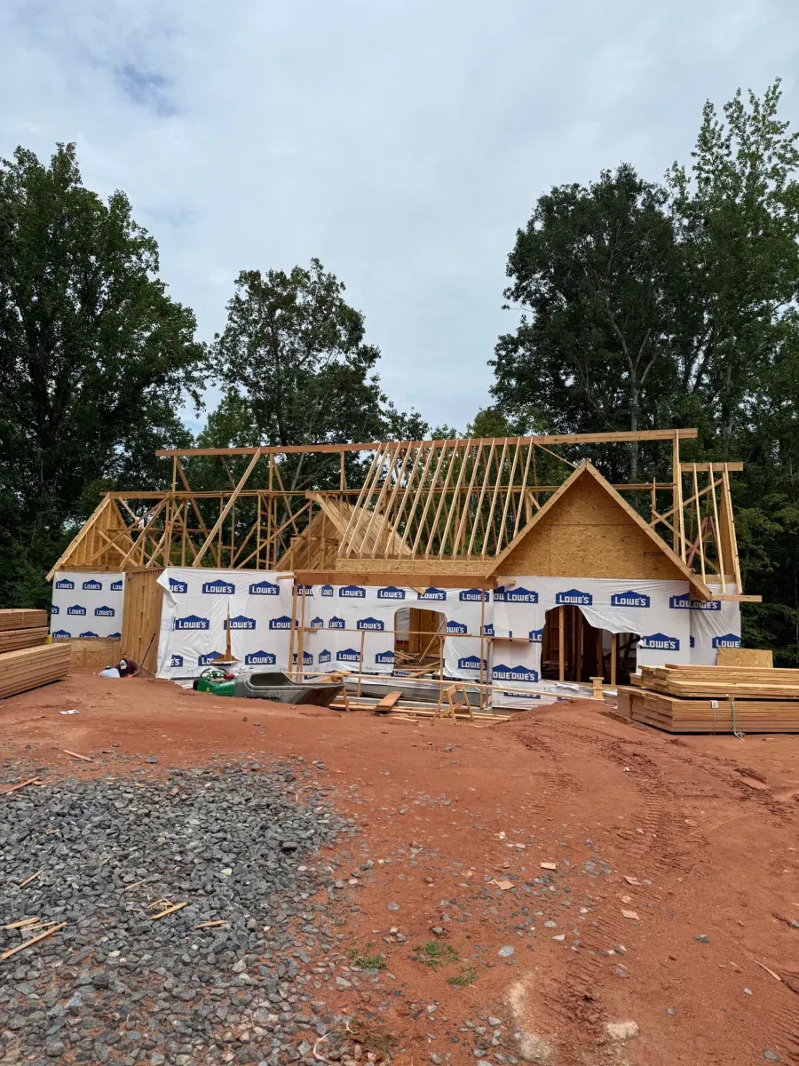 Partially built house with exposed roof framing and white wrap, on red dirt near trees