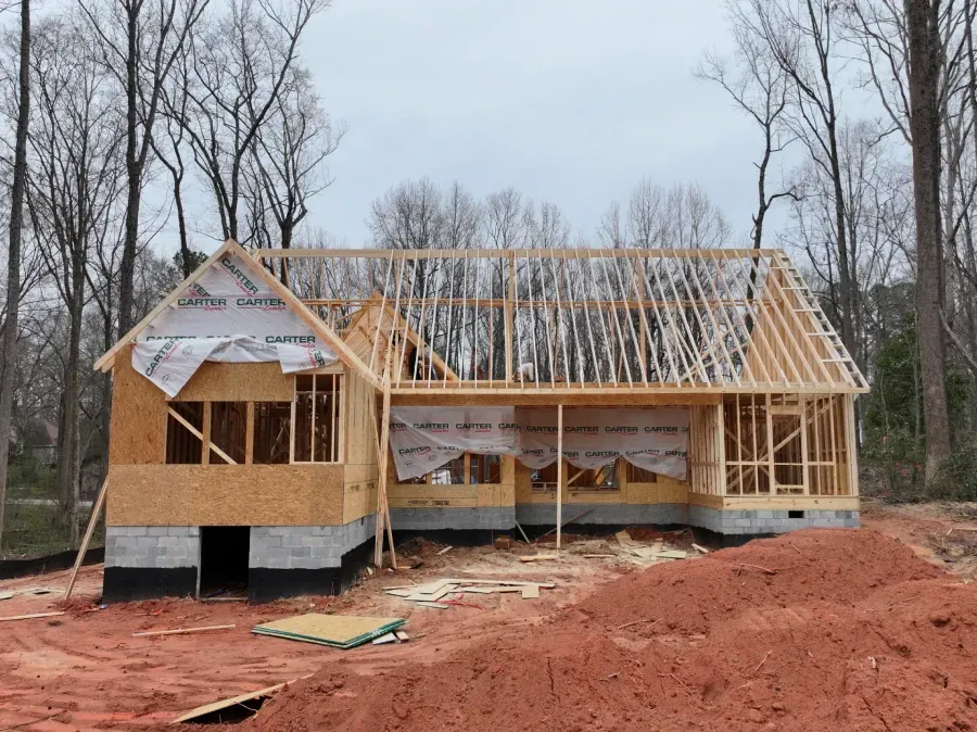 Wood-framed house under construction with roof trusses and red dirt foreground, surrounded by bare trees