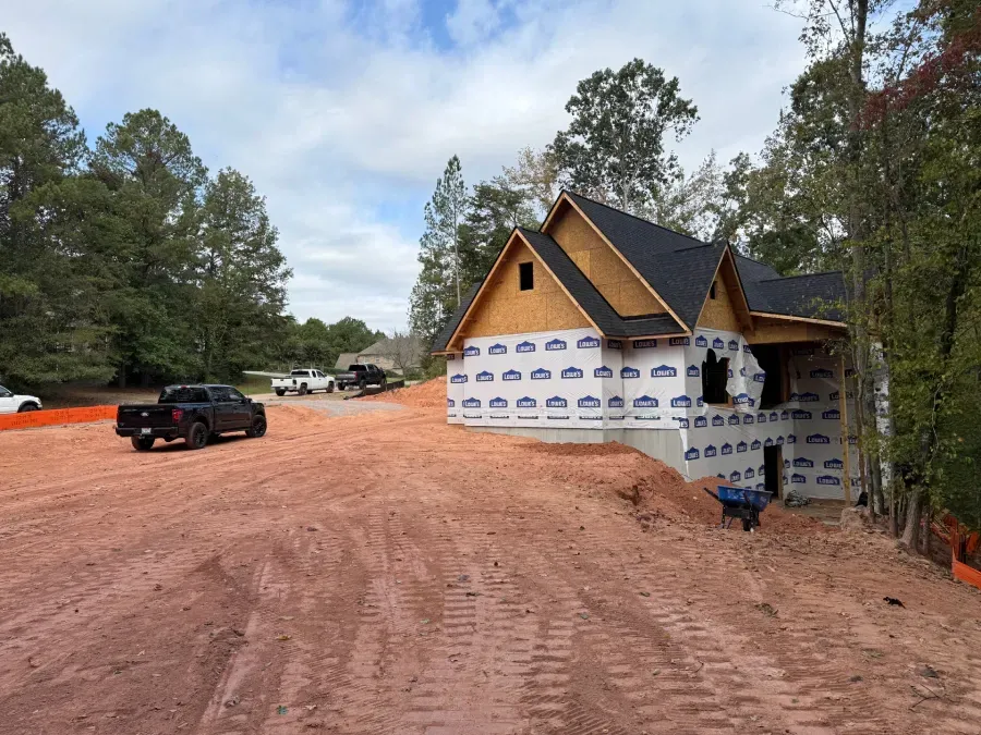 Partially built house wrapped in white sheathing beside a red dirt road and parked vehicles in a wooded area