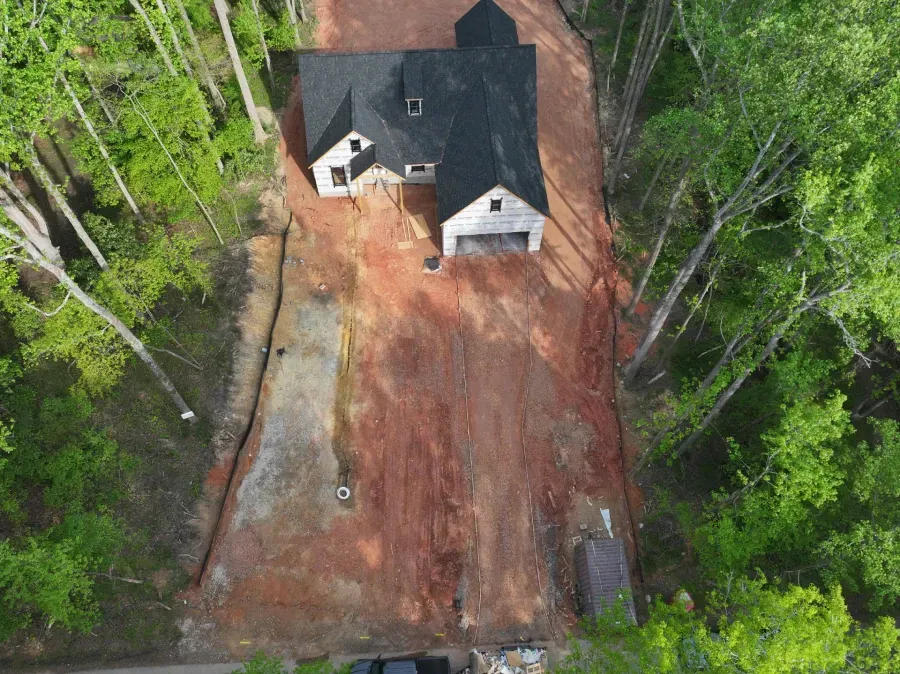 Aerial view of a house under construction with a red dirt lot and surrounding green trees.